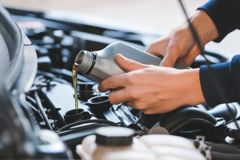 A close-up of an individual pouring golden motor oil from a silver bottle into a car engine during an oil change.