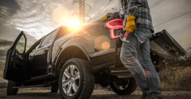 A man wearing a helmet and gloves while holding a hammer, leaning on the back side of a black pickup truck.