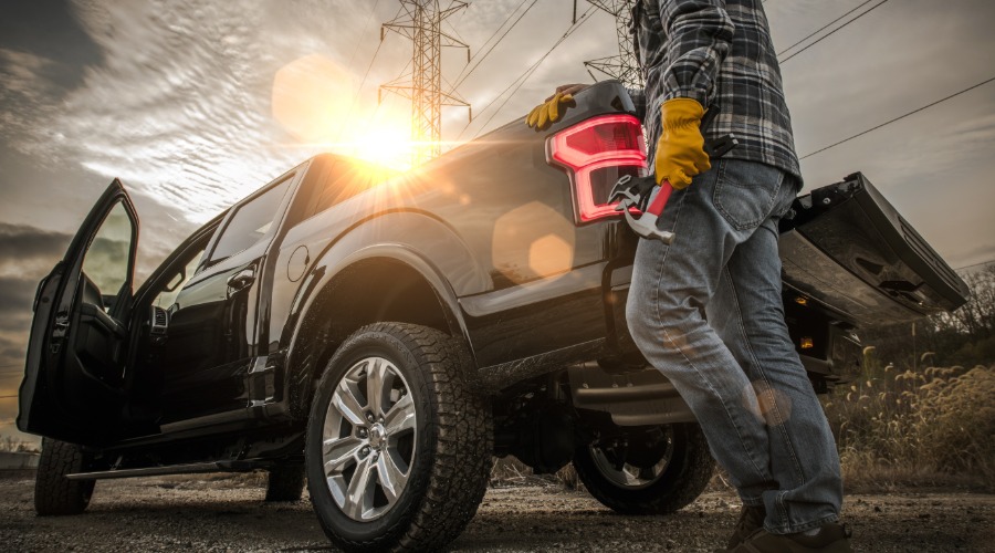 A man wearing a helmet and gloves while holding a hammer, leaning on the back side of a black pickup truck.