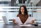 A businesswoman sits at her desk and reviews two documents. She has a laptop and a calculator at her side.