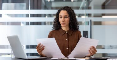 A businesswoman sits at her desk and reviews two documents. She has a laptop and a calculator at her side.