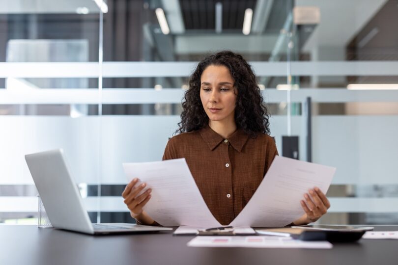 A businesswoman sits at her desk and reviews two documents. She has a laptop and a calculator at her side.