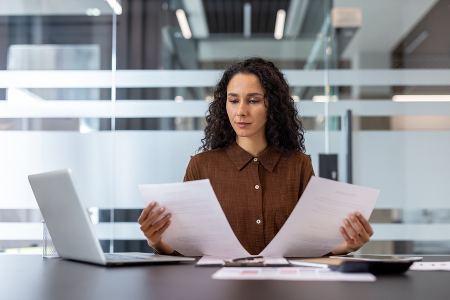 A businesswoman sits at her desk and reviews two documents. She has a laptop and a calculator at her side.