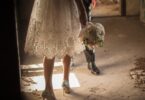 Bride in lace dress holds white bouquet, glitter heels stepping through dusty doorway; groom follows behind.