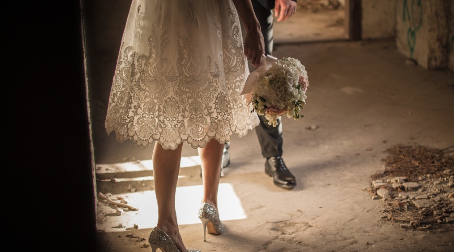 Bride in lace dress holds white bouquet, glitter heels stepping through dusty doorway; groom follows behind.