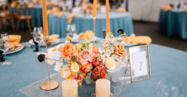 A centerpiece on a table for a rehearsal dinner. There are orange candles and an arrangement of flowers in the center.