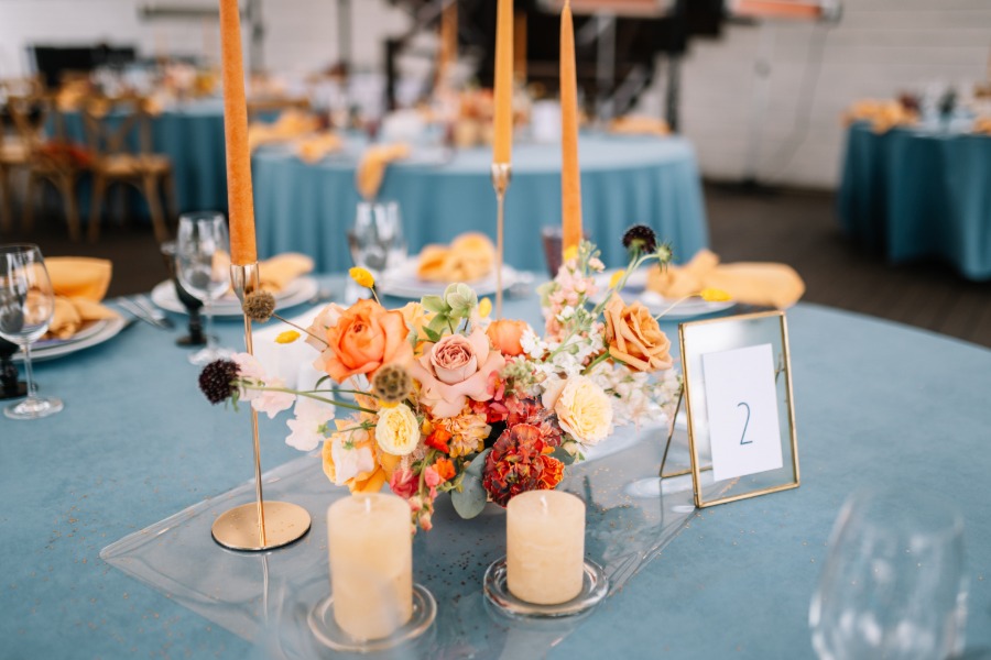 A centerpiece on a table for a rehearsal dinner. There are orange candles and an arrangement of flowers in the center.