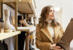 A woman in a shoe store examines a tall brown boot while standing beside shelves displaying various footwear.