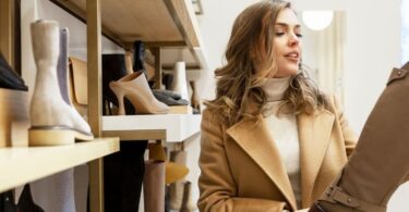 A woman in a shoe store examines a tall brown boot while standing beside shelves displaying various footwear.