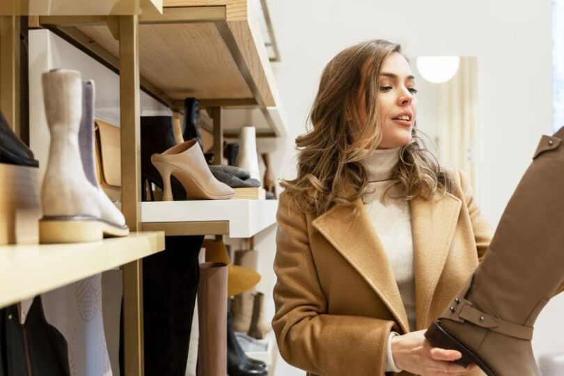 A woman in a shoe store examines a tall brown boot while standing beside shelves displaying various footwear.