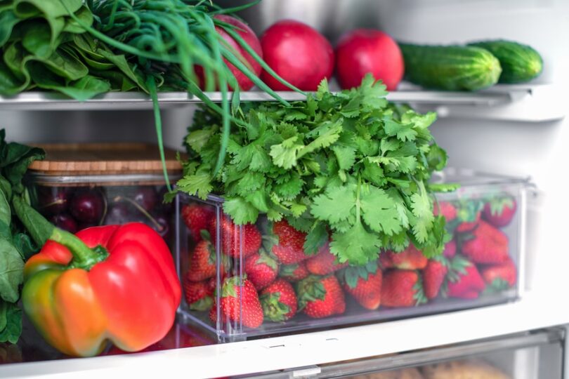 A refrigerator contains fresh produce including cilantro bunches, red bell peppers, strawberries, and cucumbers.