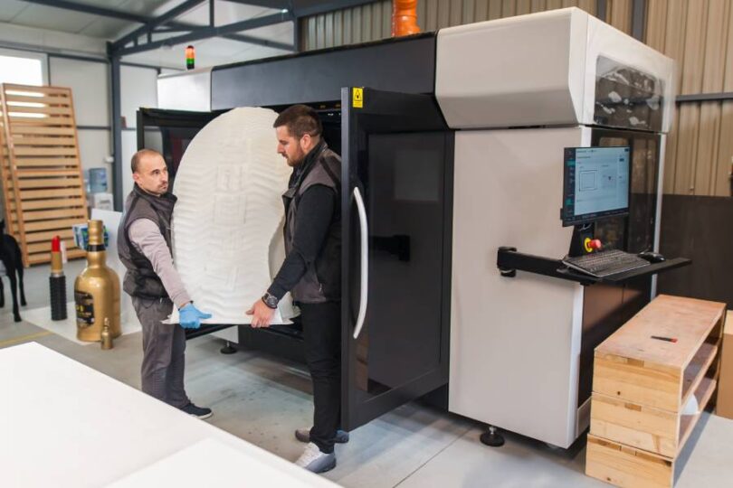 Two men carrying a massive egg-shaped 3D print out of an industrial 3D printer in a metal building.