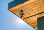 A white security camera is installed in a wooden awning. There is a blue sky in the background.
