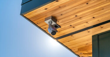 A white security camera is installed in a wooden awning. There is a blue sky in the background.