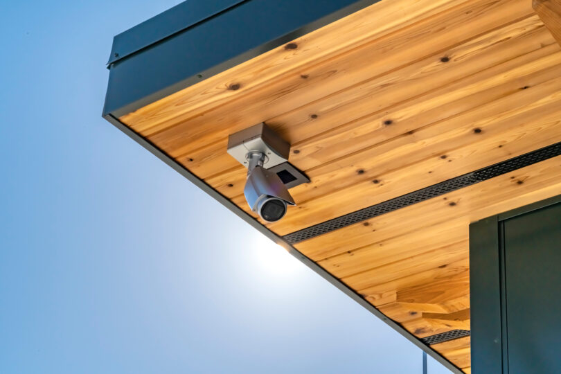 A white security camera is installed in a wooden awning. There is a blue sky in the background.