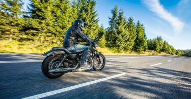 A person wearing a helmet and a leather jacket while riding a motorcycle along an empty, tree-lined road.