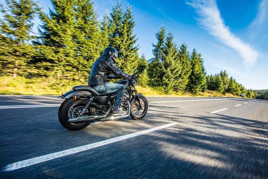A person wearing a helmet and a leather jacket while riding a motorcycle along an empty, tree-lined road.