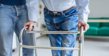 Two men standing next to each other with one of them leaning on a mobility walker. The other person is helping them.