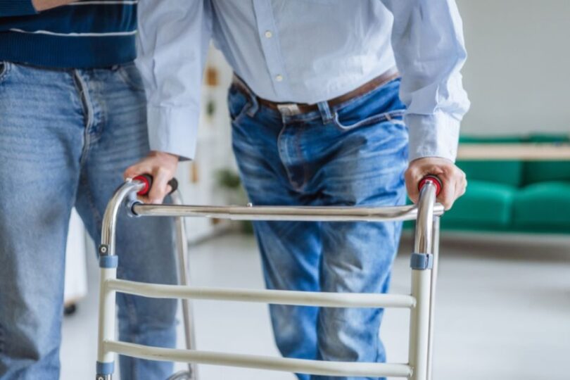 Two men standing next to each other with one of them leaning on a mobility walker. The other person is helping them.