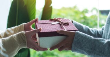 One person is handing another person a box. The box has a bow on it and there are trees in the background.