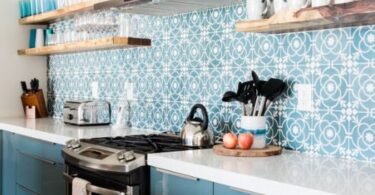 A modern kitchen with blue cabinets, tile backsplash, open shelves, stove, coffee maker, and neatly arranged utensils.