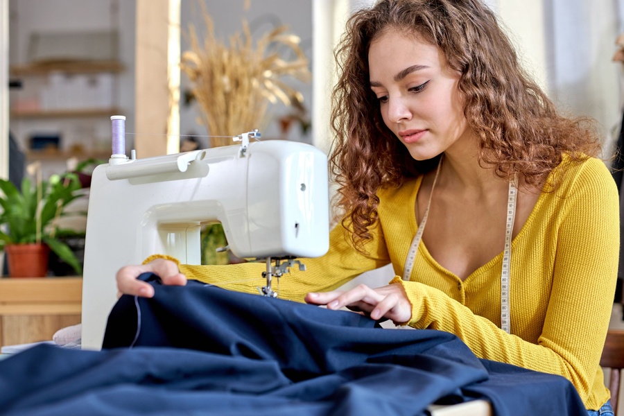 A woman guiding a large piece of navy blue fabric through a white sewing machine. She wears a tape measurer over her shoulders.