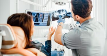 A woman is sitting in a chair while a man is holding up an x-ray of a skull. The man is pointing at the x-ray.