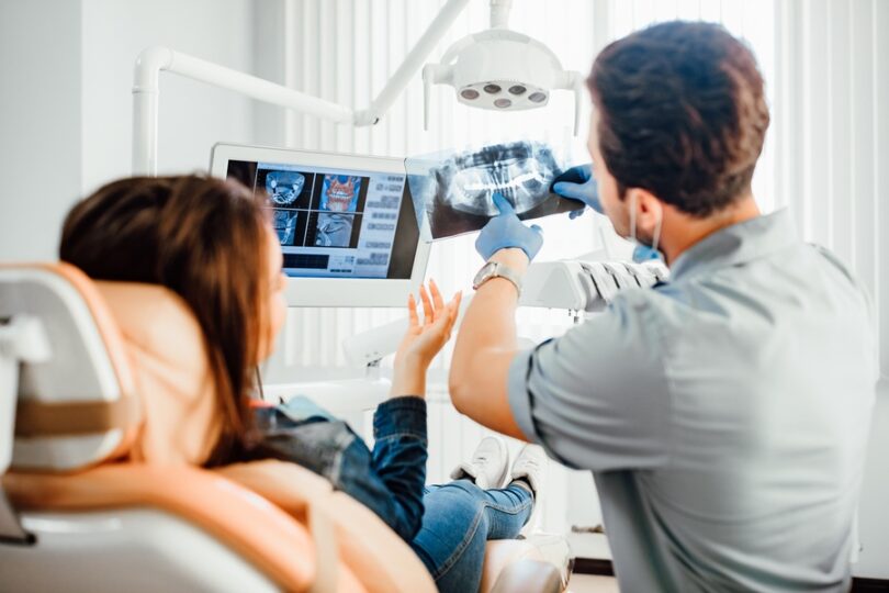 A woman is sitting in a chair while a man is holding up an x-ray of a skull. The man is pointing at the x-ray.