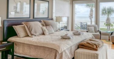 The inside of a fully decorated and furnished bedroom that has a beachside view of palm trees, sand, and water.