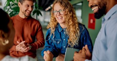 Two men are standing on either side of a woman with long, curly hair. The woman is shaking hands with someone.