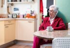 An older woman with white hair and glasses sits alone at a kitchen table, resting her hands near a white coffee mug.