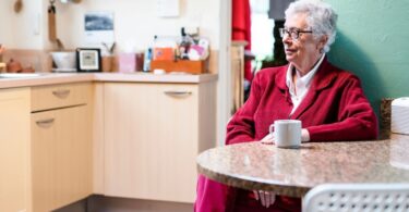 An older woman with white hair and glasses sits alone at a kitchen table, resting her hands near a white coffee mug.