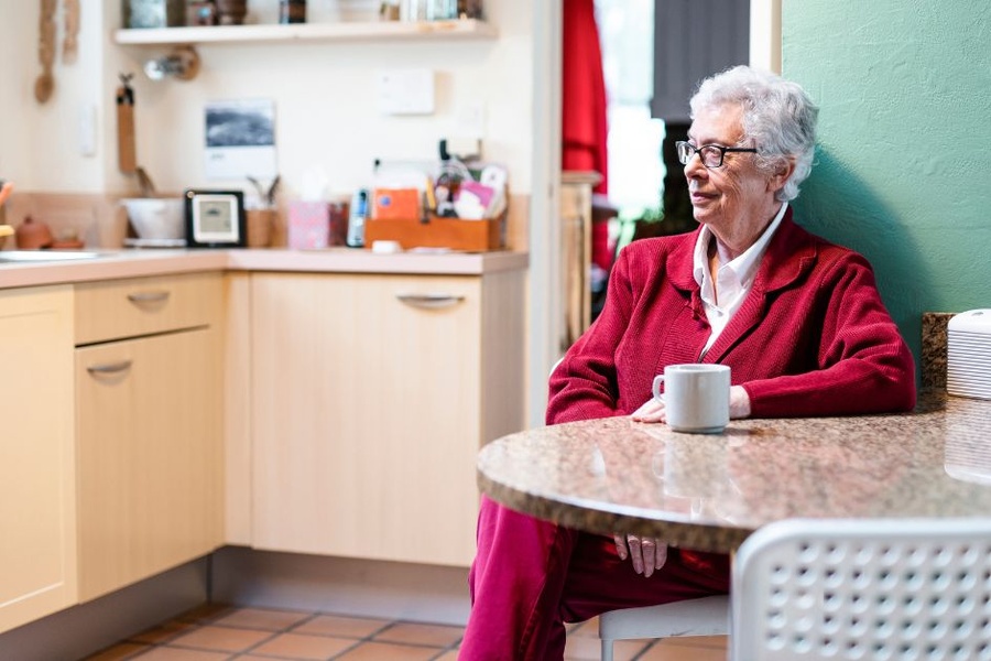 An older woman with white hair and glasses sits alone at a kitchen table, resting her hands near a white coffee mug.