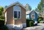 Three small colorful cottages with large front windows line a quiet path, surrounded by trees and neatly trimmed bushes.