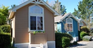 Three small colorful cottages with large front windows line a quiet path, surrounded by trees and neatly trimmed bushes.