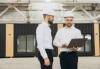 Two businessmen wearing white hard hats stand in a warehouse in front of a modular office space, using a laptop.