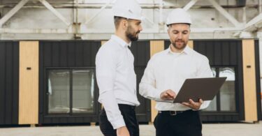Two businessmen wearing white hard hats stand in a warehouse in front of a modular office space, using a laptop.