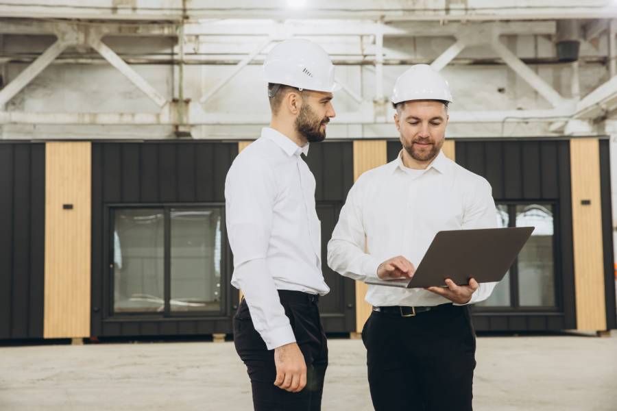 Two businessmen wearing white hard hats stand in a warehouse in front of a modular office space, using a laptop.