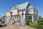 Three men on ladders working on the exterior of an old, two-story home with white siding on a sunny day.