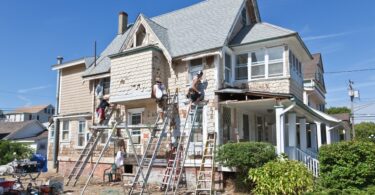 Three men on ladders working on the exterior of an old, two-story home with white siding on a sunny day.