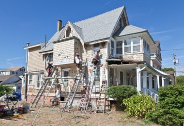 Three men on ladders working on the exterior of an old, two-story home with white siding on a sunny day.