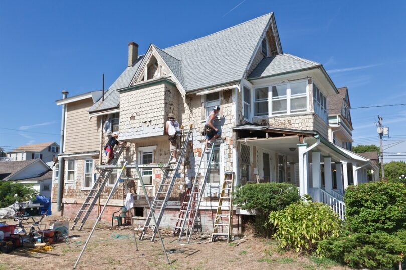 Three men on ladders working on the exterior of an old, two-story home with white siding on a sunny day.