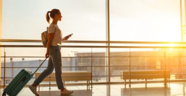 Young woman walks by an airport window hauling a suitcase, waiting to board a plane at the terminal gate.