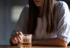 A sad woman sits at a wooden table with one arm resting on the table and the other grasping a small glass with alcohol.