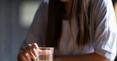 A sad woman sits at a wooden table with one arm resting on the table and the other grasping a small glass with alcohol.