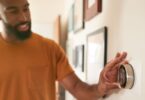 A man in an orange shirt adjusts a smart thermostat on a wall in a home hallway, smiling while setting the temperature.