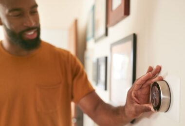 A man in an orange shirt adjusts a smart thermostat on a wall in a home hallway, smiling while setting the temperature.