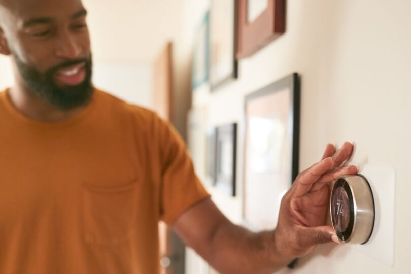 A man in an orange shirt adjusts a smart thermostat on a wall in a home hallway, smiling while setting the temperature.
