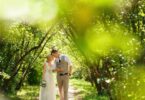 A bride and groom walking hand in hand through a lush park path, both lean into one another with their foreheads touching.