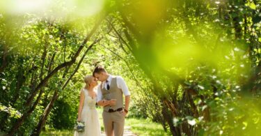 A bride and groom walking hand in hand through a lush park path, both lean into one another with their foreheads touching.